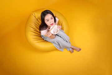 Wall Mural - Beautiful young south east Asian woman holding empty chopstick soup spoon bowl posing see eat taste smell feed offer satisfaction yummy siting on beanbag seat yellow orange background top view