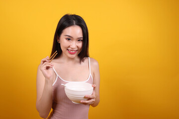 Beautiful young south east Asian woman holding chines empty chopstick Chinese soup spoon bowl utensil pretend acting posing see eat taste smell feed offer satisfaction yummy yellow orange background