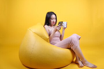 Wall Mural - Beautiful young south east Asian woman sit relax on a yellow orange beanbag seat color background hold white coffee cup smart phone