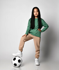 Active Asian kid girl in green sportswear and white sneakers stands with classic soccer football ball under her foot on gray background. Happy Korean soccer player kid with big smile laughing