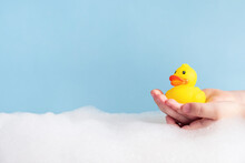 Child Hands Holding Yellow Rubber Duck In Bubble Bath On Pastel Blue Background. Baby Hygiene, Accessories For Bathing. Copy Space