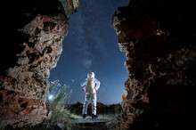 Back View Of Spaceman In White Suit And Helmet Is Standing Between Abandoned Walls At Night. Man In Spacesuit Is Looking In Starry Sky In Isolated Area. Concept Of Cosmic Traveler.