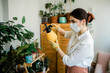 © Beznika - woman spraying water on a house plant and flower with a spray bottle at home. Uses insecticide, mask and gloves