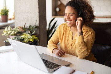 Smiling multiracial woman with Afro hairstyle wearing headset using laptop computer for video connection, consults client online, working remotely from home office