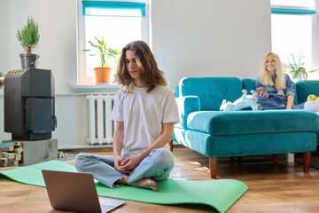 Wall Mural - Guy teenager sitting in lotus position on yoga mat at home on floor with laptop