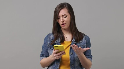 Wall Mural - Sad young woman 20s years old in denim jacket yellow t-shirt using mobile cell phone swear show thumb down hears fake news unexpected rumor has some problems isolated on grey color background studio