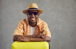 © Studio Romantic - Cheerful handsome African American man with toothy smile going on summer vacation trip. Studio portrait of happy young black tourist in sunglasses and sun hat with yellow travel suitcase