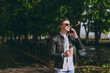 © ViDi Studio - Young friendly stylish man in black leather jacket eyeglasses walk in green park hold takeaway craft paper cup drink coffee to go talk by mobile cell phone Concept of urban lifestyle spring season