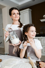 A beautiful woman in whose hands a can of flour and a little girl in aprons are standing in the kitchen. Family cooking