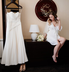 Wall Mural - bride in a white silk robe sits on the table near the wedding dress on a hanger.