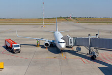 White Airplane Docked On Apron Outside Airport Terminal With Jet Bridge. Worker Refuels Plane. Fuel Hose From Red Truck Connected Under The Fuel Tank In The Wing. Ground Service Theme.