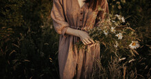 Beautiful Woman In Linen Dress Gathering Wildflowers In Summer Meadow In Evening.  Stylish Young Female In Rustic Dress Picking Flowers In Countryside. Atmospheric Stylish Vintage Image
