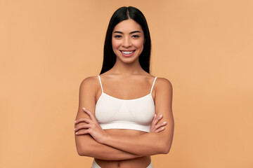 Wall Mural - Young Asian woman in white lingerie crossed her arms on a beige background. Sports, healthy lifestyle. Spa body care. Plastic surgery. Cosmetology concept.