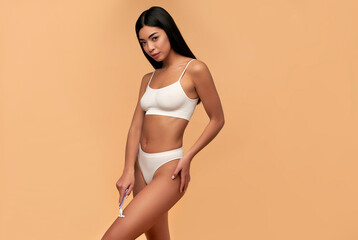 Wall Mural - Young Asian woman in white lingerie with a straight razor on a beige background. Depilation, body care.