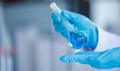 Wall Mural - Close up hand laboratory beaker with blue liqid in analyst's hand in plastic glove blur background. Concept Science experiment.