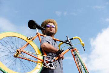 Hispanic hipster carrying a colorful fixie bicycle on his shoulder with a blue sky as background. Sustainable mobility and transport concept.