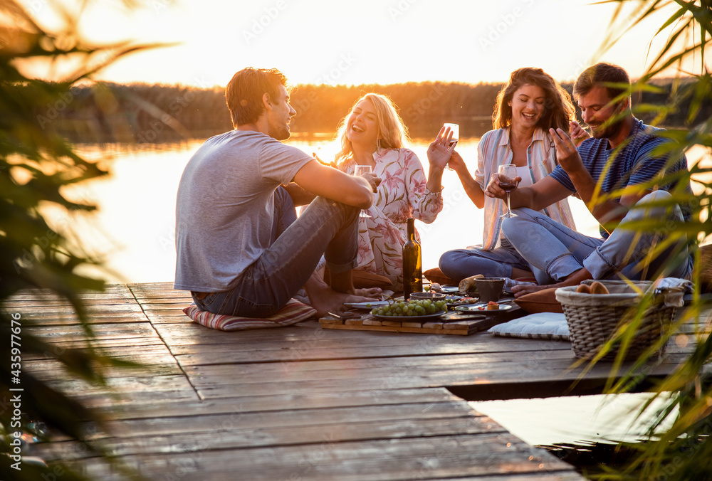 Group of friends having fun on picnic near a lake, sitting on pier eating and drinking wine. - obrazy, fototapety, plakaty 