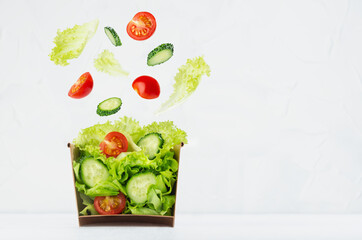 Fresh vegetable salad in craft box for take away food with slices of ingredients levitate - cherry tomato, cucumber, green salad on white wood table, copy space.