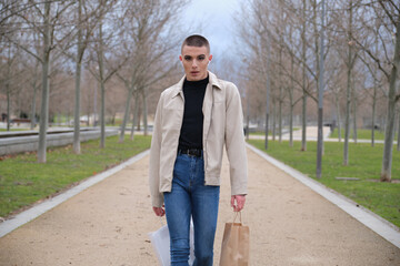 Wall Mural - Young man wearing make up walking with shopping bags and listening music in a park. Queer person.