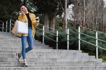 Wall Mural - Young non binary person laughing and speaking on the smartphone going down stairs with shopping bags and a coffee cup. Make up androgynous person.