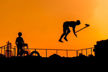 Wall Mural - Unrecognizable teenage boy silhouette showing high jump tricks on scooter against orange sunsetwarm sky at skatepark. Sport, extreme, freestyle, outdoor activity concept