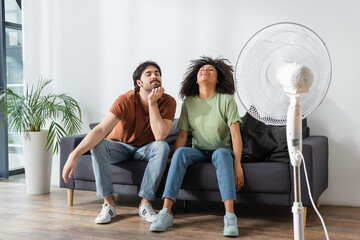 cheerful interracial man and woman sitting on couch near blurred electric fan in living room