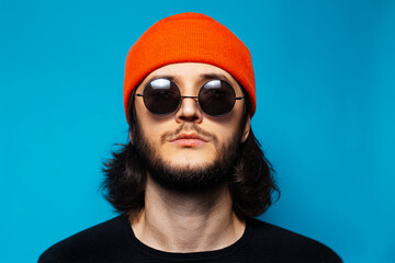 Wall Mural - Studio portrait of young confident man on blue background. Guy with long hair wearing orange hat, round sunglasses and black sweater; looking up.