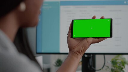 Black woman looking at isolated display having online videocall conference meeting using mock up green screen chroma key phone sitting at desk in living room. Student watching social media video