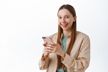 Wall Mural - Smiling young attractive girl using mobile phone, chatting on smartphone and looking at camera pleased, standing over white background