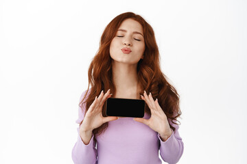 Wall Mural - Glamour shopping girl with ginger hair, kissing and showing horizontal screen of mobile phone, display an app on smartphone, standing against white background