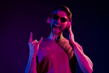 Sticker - Portrait of attractive cheerful crazy guy listening bass sound showing horn sing fooling isolated over dark neon violet color background