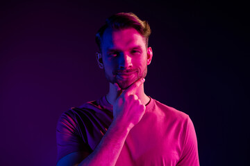 Portrait of attractive cheerful well-groomed guy touching chin isolated over dark neon light violet color background