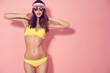 © zamuruev - Beautiful young female in cap, sunglasses and yellow swimsuit looking away while standing on pink background.