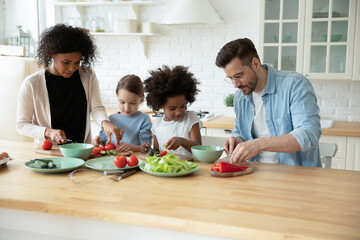 Loving young multiracial family with diverse small daughters cook healthy vegetarian salad for dinner lunch in kitchen. Caring multiethnic mom and dad prepare food with children. Adoption concept.