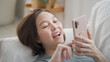© Vajirawich - happy asian woman face relaxing using mobile smart phone surfing internet and chat on sofa couch at home. female laying down in living room embarrassed smile when using mobile phone. woman laugh happy