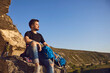 © Studio Romantic - Active tourism. Dreamy young hiker man sitting on the edge of the mountain and looking into the distance. Side view of a guy who sat down to relax while traveling on a mountainous rocky terrain.