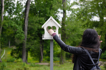 Wall Mural - girl feeds birds in a feeder in an spring park, back view. girl puts food. taking care of birds, natural green background, bokeh, focus. wooden birdhouse. white bird feeder and woman in the garden