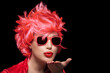 Stylish young woman wearing trendy modern sunglasses and red wig blowing across the palm of her hand sending a kiss