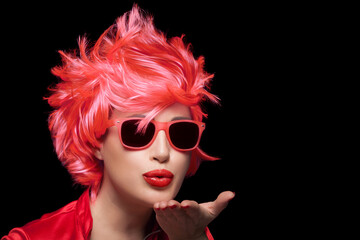 Wall Mural - Stylish young woman wearing trendy modern sunglasses and red wig blowing across the palm of her hand sending a kiss