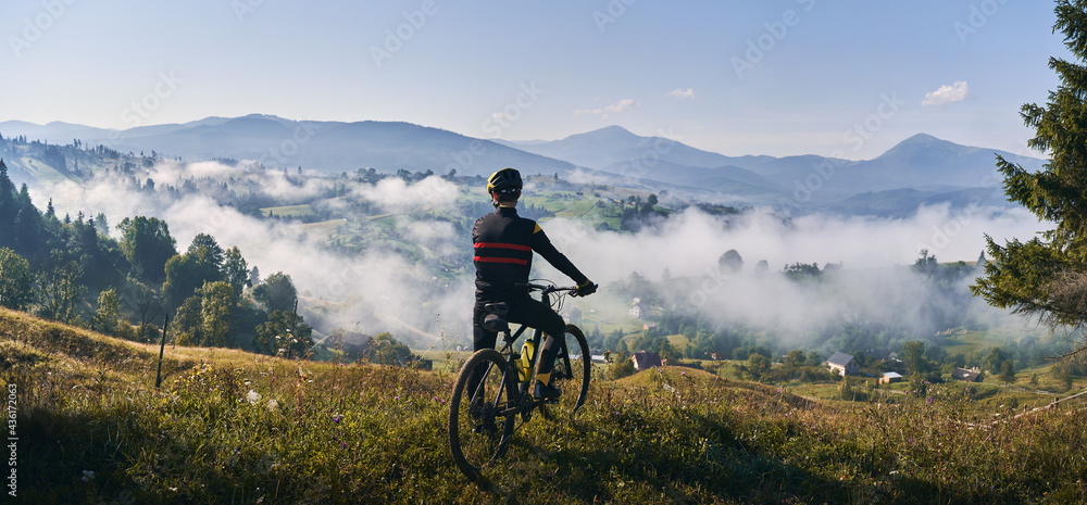 Man riding bicycle on grassy hill and looking at beautiful misty mountains. Male bicyclist enjoying panoramic view of majestic mountains during bicycle ride. Concept of sport, bicycling and nature. - obrazy, fototapety, plakaty 