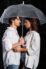 Wall Mural - side view of romantic young adult boyfriend and girlfriend standing in rain with umbrella on black background