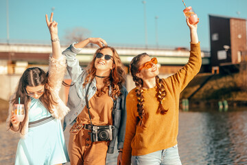 Wall Mural - Three girls near the river against the sky having fun. Fashion girls in sunglasses. Jump, rejoice, drink drinks, smile have fun, going crazy. Color dressed friends spend summer vibes, dance