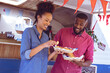 © Wavebreak Media - Smiling diverse couple eating hot dogs and potato wedges by food truck on sunny day