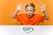 © Roman - a boy in an orange T-shirt sits at a white table and emotionally raises his hands and looks at the work done with thin paper strips in the quilling technique