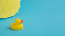 Yellow Rubber Duck With Umbrella On A Blue Background. Summer Vacation Trip. Minimal Concept.