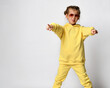 Portrait of an active funny little girl pointing fingers at you. Cheerful girl dressed in a yellow sports suit and sunglasses shows her tongue while standing on a white background.