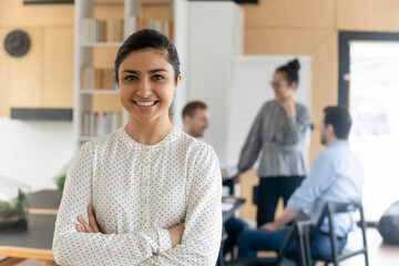 Poster - Head shot portrait smiling confident Indian businesswoman with arms crossed standing in modern office with colleagues on background, successful happy employee entrepreneur executive looking at camera