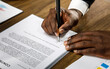 © Bangkok Click Studio - Close up and selective focus on African black formal professional business male hands holding pen, signing in white paper form or application to confirm and deal contract agreement on table.