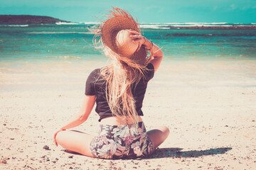 Wall Mural - Summer lifestyle blonde long hair young woman sitting at the beach on the sand enjoying blue ocean water in background holding straw hat - people travel lifestyle and holiday vacation outdoor