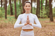 Young adult concentrated female wearing white sports top keeping palms together, looking directly at camera, doing yoga in open fresh air in green forest.